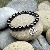 Black Agate Pentacle Charm Bracelet displayed on a bed of river rocks, showcasing its glossy black gemstone beads.