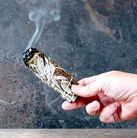 Hand holding a lit Sage and Basil Herb Bundle, with one end smoldering, and a trickle of smoke rising upward from the end.