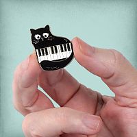 The Piano Cat Enamel Pin held in someone's fingertips, showing its small size, and its black, white, and gold colors.