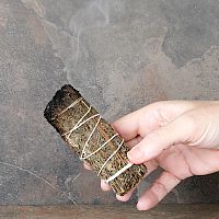 Hand holding a lit Mugwort Herb Bundle, with one end smoldering, and a trickle of smoke rising upward from the end.