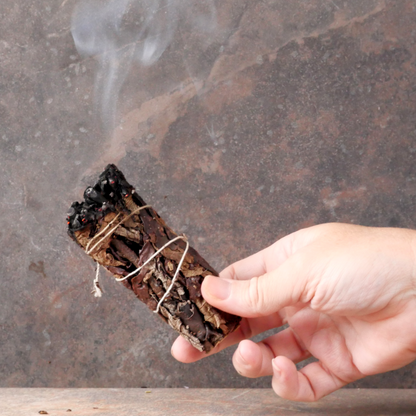 Hand holding a lit Yerba Santa Herb Bundle, with one end smoldering, and a trickle of smoke rising upward from the end.