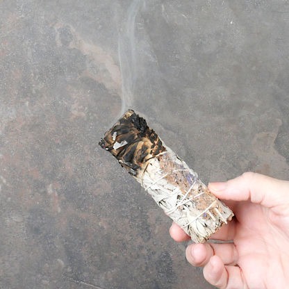 Hand holding a lit Sage and Lavender Herb Bundle, with one end smoldering, and a trickle of smoke rising upward from the end.