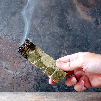 Hand holding a lit Sage and Cedar Herb Bundle, with one end smoldering, and a trickle of smoke rising upward from the end.